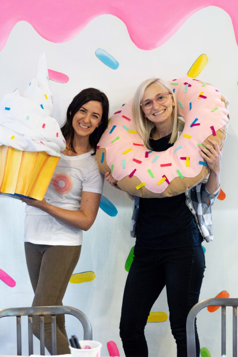 Rory and Volta in front of the sprinkled donut wall holding cupcake and donut props